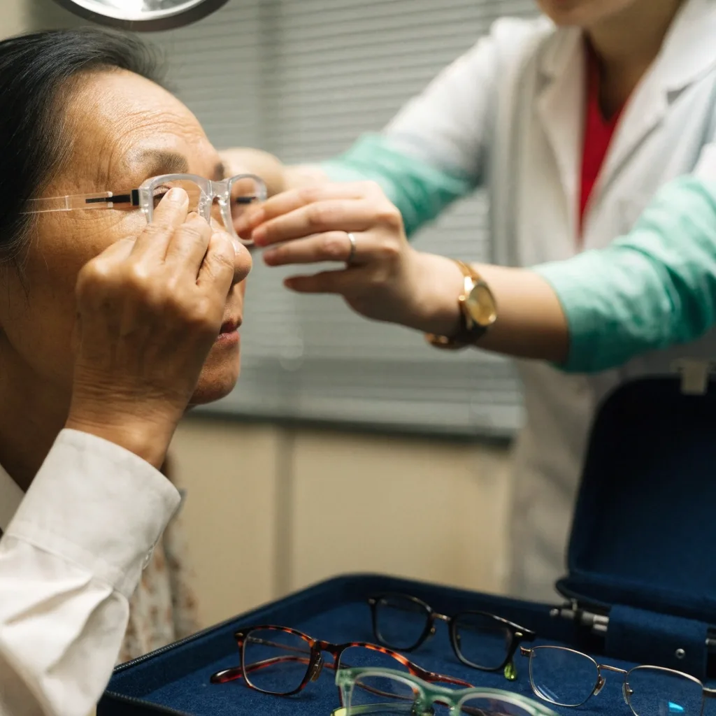 An older woman trying on trial glasses at an optical fitting table with several frames laid out in front of her