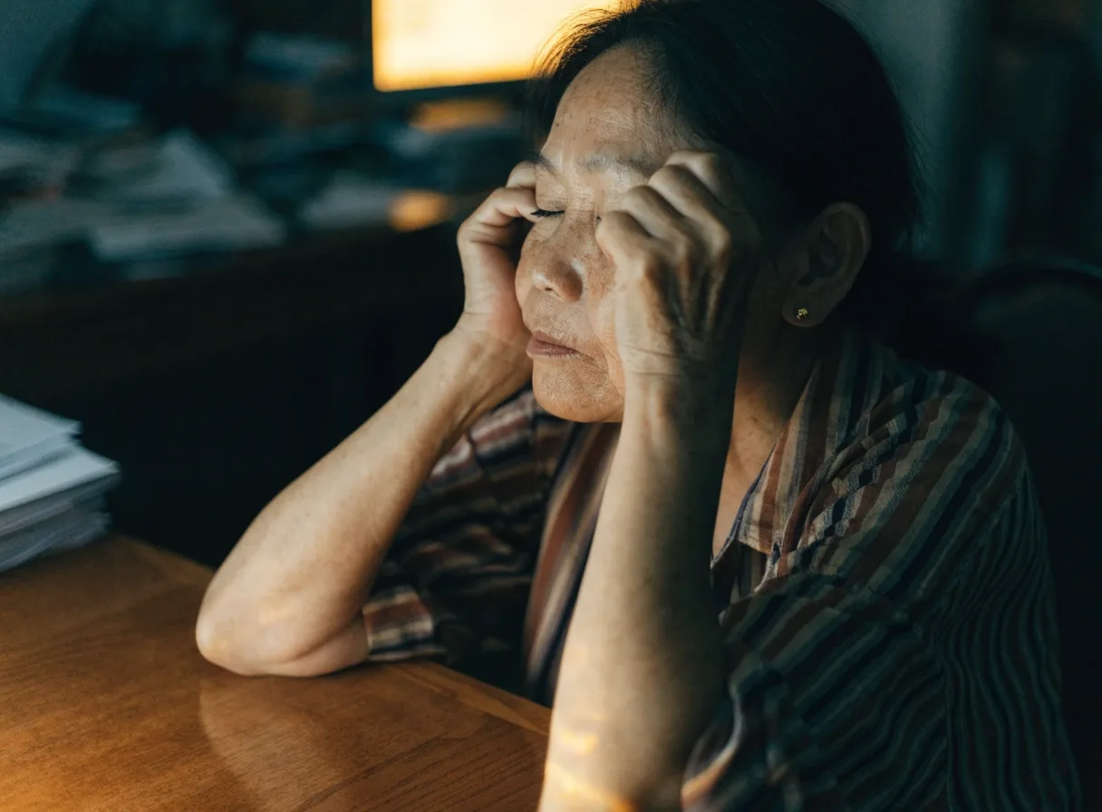 An older woman at a desk in the evening pressing two fingers gently against her closed eyelids
